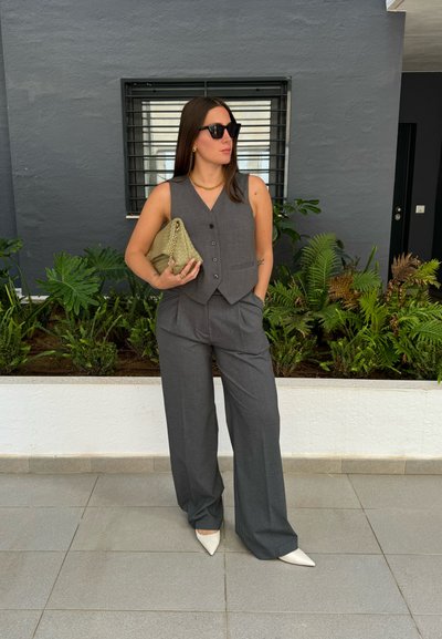 Chaleco gris a medida y pantalones de pierna ancha, combinados con tacones de punta blanca. Sosteniendo un bolso de mano de textura verde oliva. Plantas verdes en el fondo.
