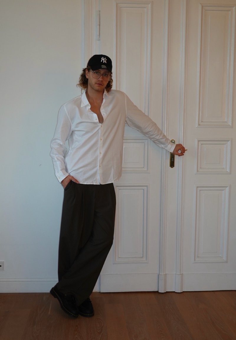 Un homme portant une chemise blanche, un pantalon noir et une casquette noire se tient appuyé contre une porte blanche fermée, une main dans la poche.