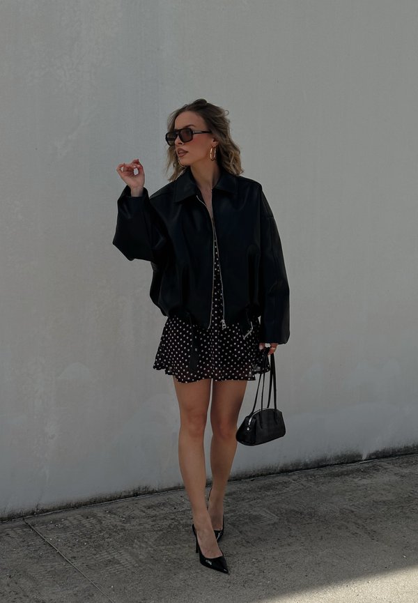 Femme portant une veste en cuir noire sur une robe à pois noire et blanche, des talons hauts noirs, des lunettes de soleil et portant un petit sac à main noir.