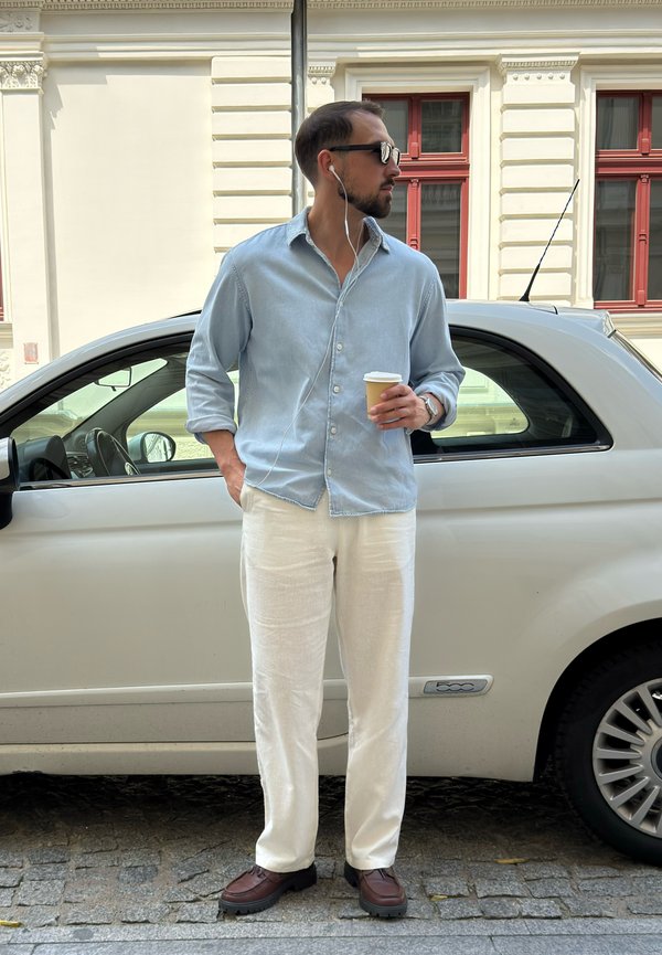 Homme portant des lunettes de soleil et des écouteurs se tient à côté d'une voiture blanche, tenant une tasse de café, habillé d'une chemise bleu clair et d'un pantalon blanc.