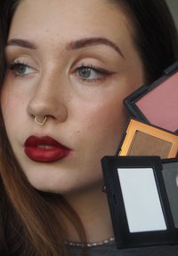 Fyra kosmetikpaletter hålls nära en persons ansikte och visar nyanser av rosa, brunt och vitt. Personen har mörkt hår och en gyllen septumpiercing.