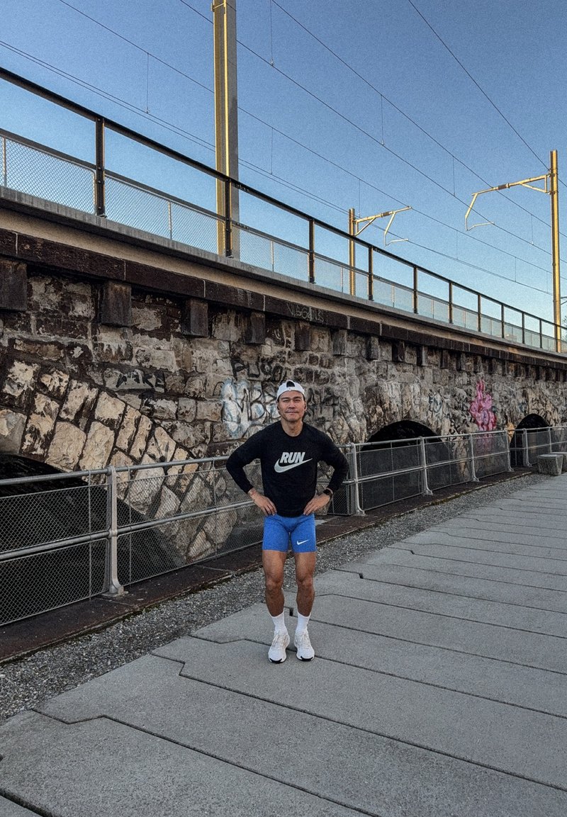 Corridore con maglia nera a maniche lunghe e pantaloncini blu in piedi su un sentiero asfaltato vicino a un muro di pietra coperto di graffiti sotto i binari del treno al crepuscolo.