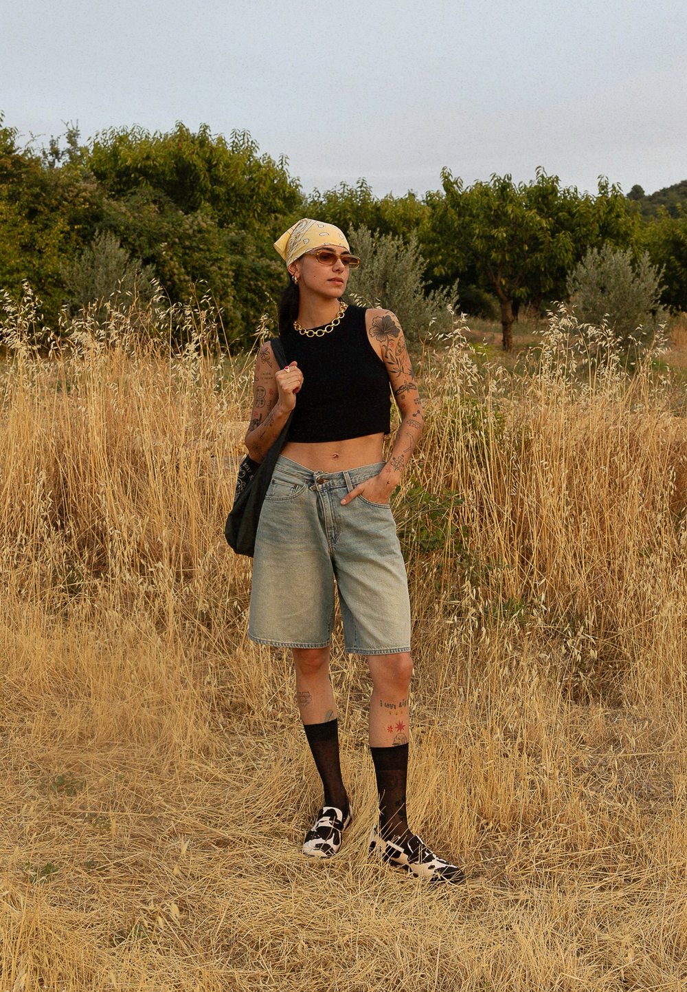 Top scurt negru, pantaloni scurți din denim albastru deschis, ciorapi din plasă negri, pantofi cu imprimeu de vacă, batic galben, colier din lanț auriu, diverse tatuaje.
