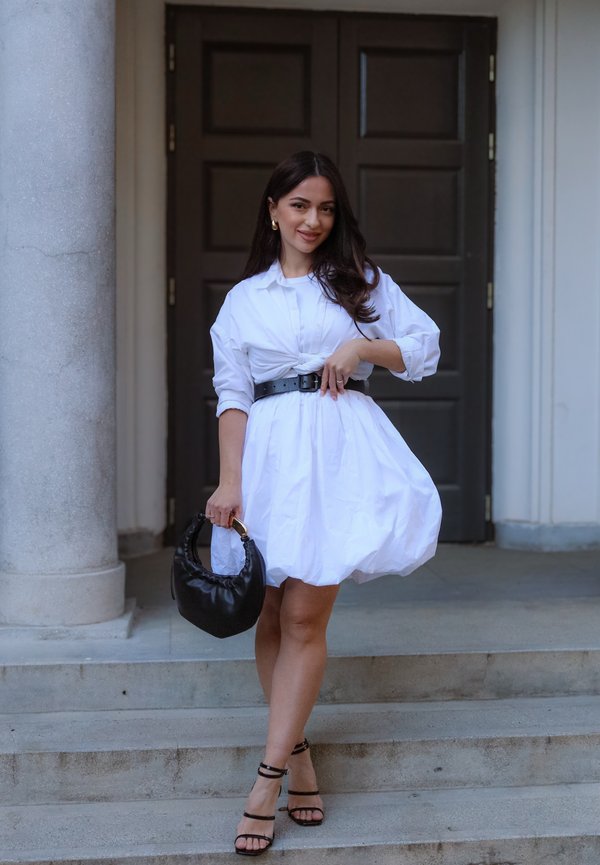 Woman standing on stone steps wearing a white dress with black belt and sandals, holding a black handbag, with columns and dark door behind her.