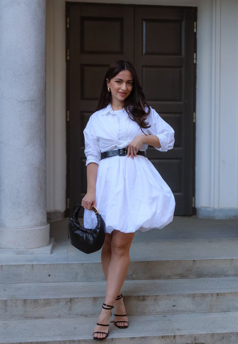 Woman standing on stone steps wearing a white dress with black belt and sandals, holding a black handbag, with columns and dark door behind her.