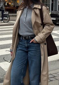Beige trench coat worn over a grey button-up knit top, matched with blue wide-leg jeans. Dark brown handbag and holding sunglasses.