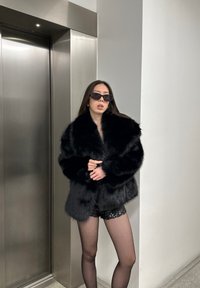 Manteau en fausse fourrure noire avec un large col, porté sur un short en sequins métalliques. Porté avec des lunettes de soleil foncées, debout près d'un ascenseur en métal.