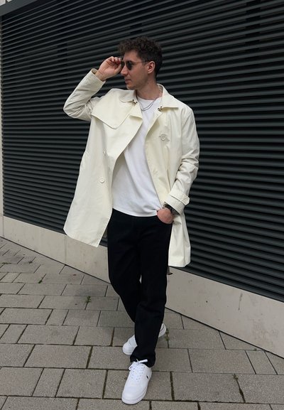 Abrigo de trench blanco y sobredimensionado con gran solapa y botones dobles, llevado sobre una camiseta blanca y jeans negros. Zapatillas blancas.