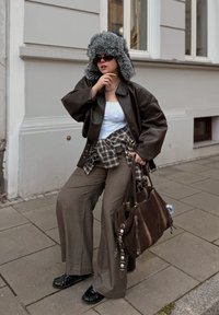 Giacca di pelle marrone sopra una canottiera bianca, camicia a quadri annodata in vita, pantaloni di lana grigia, scarpe nere e cappello di pelliccia grigia. Borsa marrone con dettagli.