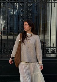 Pulover bej tricotat, fustă albă transparentă în straturi, geantă maro din piele întoarsă cu accesorii în formă de inimă, coliere suprapuse și accente aurii.