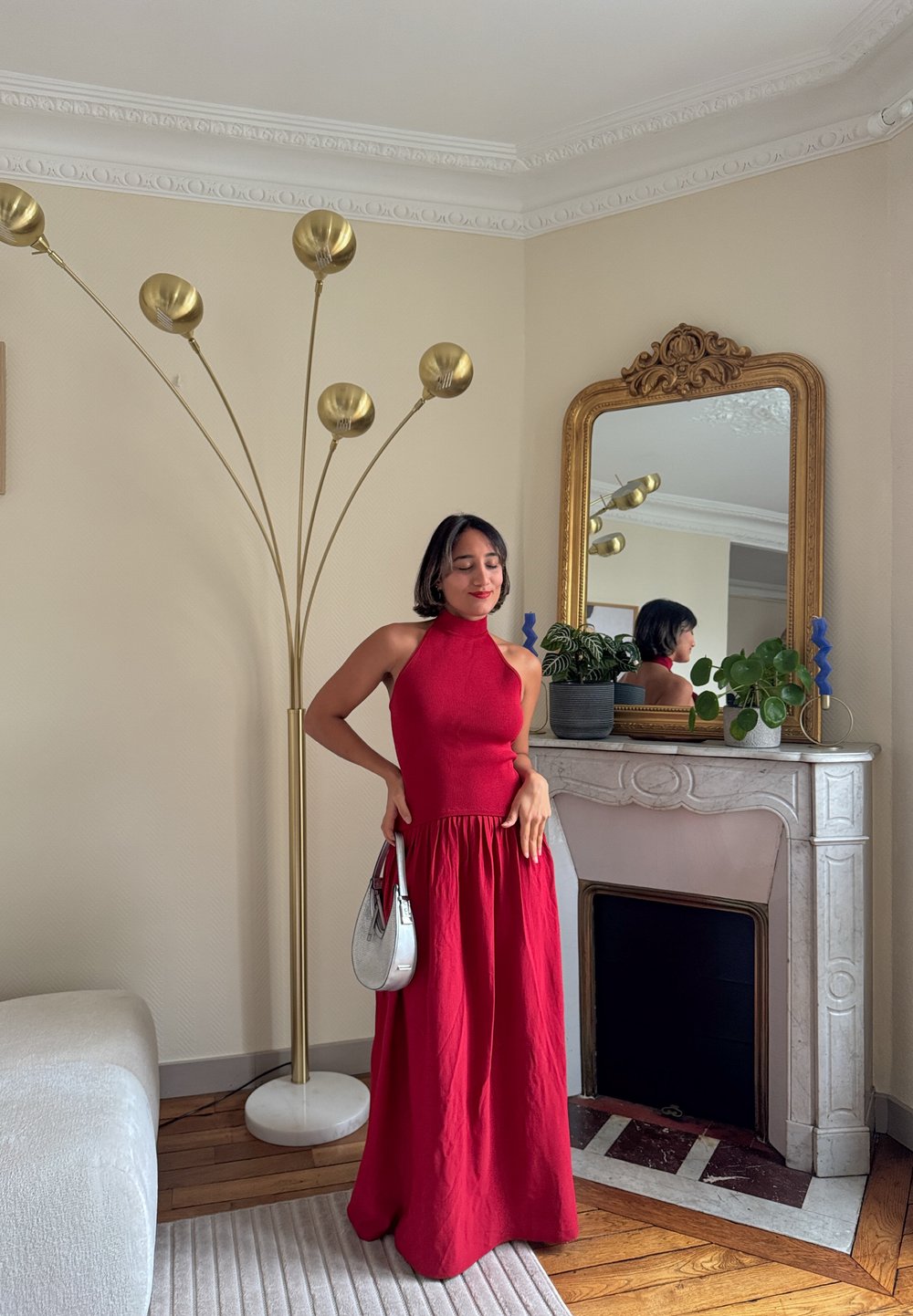 Robe maxi rouge à col licou avec un corsage ajusté et une jupe fluide. Tissu lisse et sac à main argenté brillant. Lampadaire doré en arrière-plan.