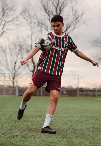 Joven jugador de fútbol masculino con camiseta de rayas y pantalones cortos color granate controla un balón de fútbol en el aire en un campo de césped con árboles sin hojas de fondo.