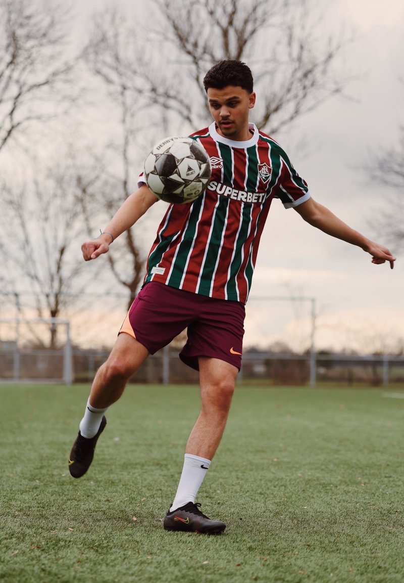 Jeune joueur de football masculin en maillot rayé et short bordeaux contrôle un ballon de football en l'air sur un terrain herbeux avec des arbres nus en arrière-plan.