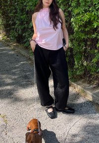 Top rosa claro, sem mangas, com decote franzido, combinado com calças negras folgadas. Sapatos pretos. Pequeno cão castanho com trela ao lado.