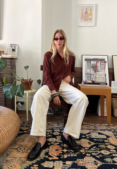 Camisa marrón de botones, pantalones anchos blancos, mocasines negros y gafas de sol oscuras pequeñas. Ambiente interior con una planta decorativa y obras de arte.