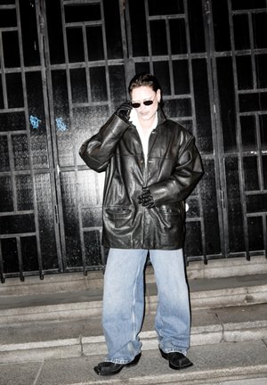 Persona con chaqueta de cuero negra extragrande, guantes negros, gafas de sol y vaqueros anchos azul claro, de pie en unos escalones de piedra frente a unas puertas metálicas negras.