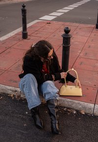 Geantă galbenă cu design structurat, mâner curbat și detaliu cu logo. Modelul poartă o jachetă neagră, blugi albaștri și cizme negre până la genunchi.