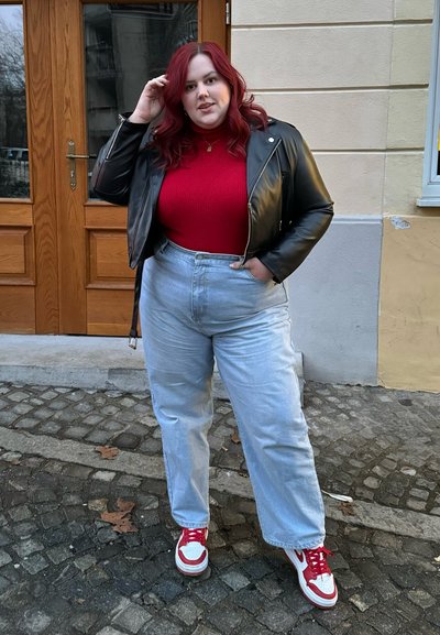 Chaqueta de cuero negra estilo motero, suéter de cuello alto acanalado rojo, jeans de tiro alto de color azul claro y zapatillas rojas y blancas. De pie sobre un camino de adoquines.