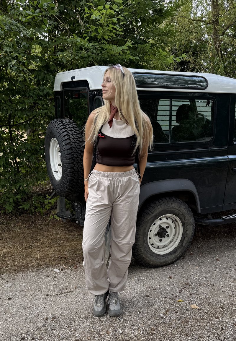 Jeune femme aux longs cheveux blonds se tient à côté d'un véhicule tout-terrain noir et blanc, portant un haut sans manches et un pantalon clair.