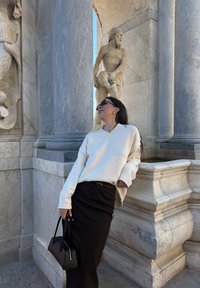 Femme en pull blanc et jupe noire s'appuie contre un détail architectural en marbre, avec une sculpture classique en pierre et des colonnes en arrière-plan.