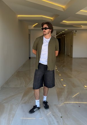 Hombre con gafas de sol, camisa verde de manga corta, camiseta blanca, pantalones cortos negros, calcetines blancos y deportivas negras de pie en un pasillo moderno.