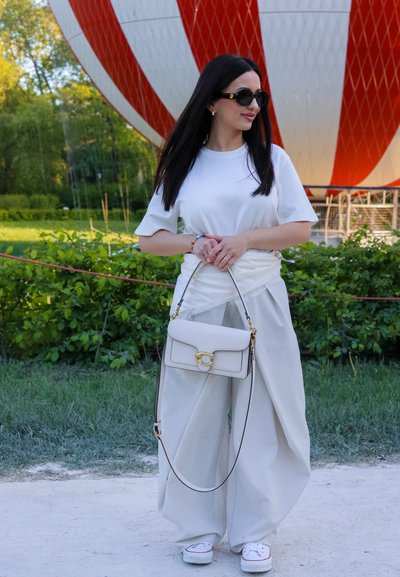 Mujer con pelo largo y oscuro que lleva gafas de sol, camisa blanca, pantalones anchos blancos y zapatillas deportivas, sosteniendo un bolso blanco cerca de un globo aerostático rojo y blanco.