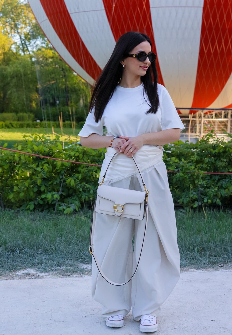Femme aux longs cheveux foncés portant des lunettes de soleil, une chemise blanche, un pantalon blanc large et des baskets, tenant un sac à main blanc près d'une montgolfière rouge et blanche.