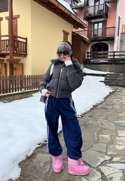 Chaqueta puffer gris con ribete de piel sintética, pantalones deportivos azul marino con rayas blancas, botas de invierno rosas con marca, fondo de camino de nieve y piedra.