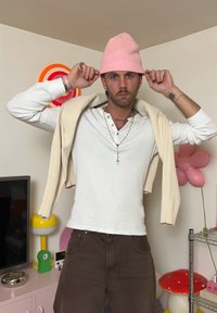 Witte langemouwen shirt, beige cardigan over de schouders gedrapeerd, roze gebreide beanie, brown pants, accessoires omvatten kettingen en ringen.
