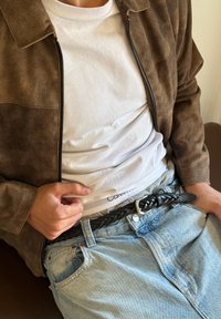 Brūna zamšāda ar rāvējslēdzēju jaka virs baltā T-krekla. Melna pītā ādas josta ar sudraba sprādzi, valkāta ar gaiši ziliem džinsiem.