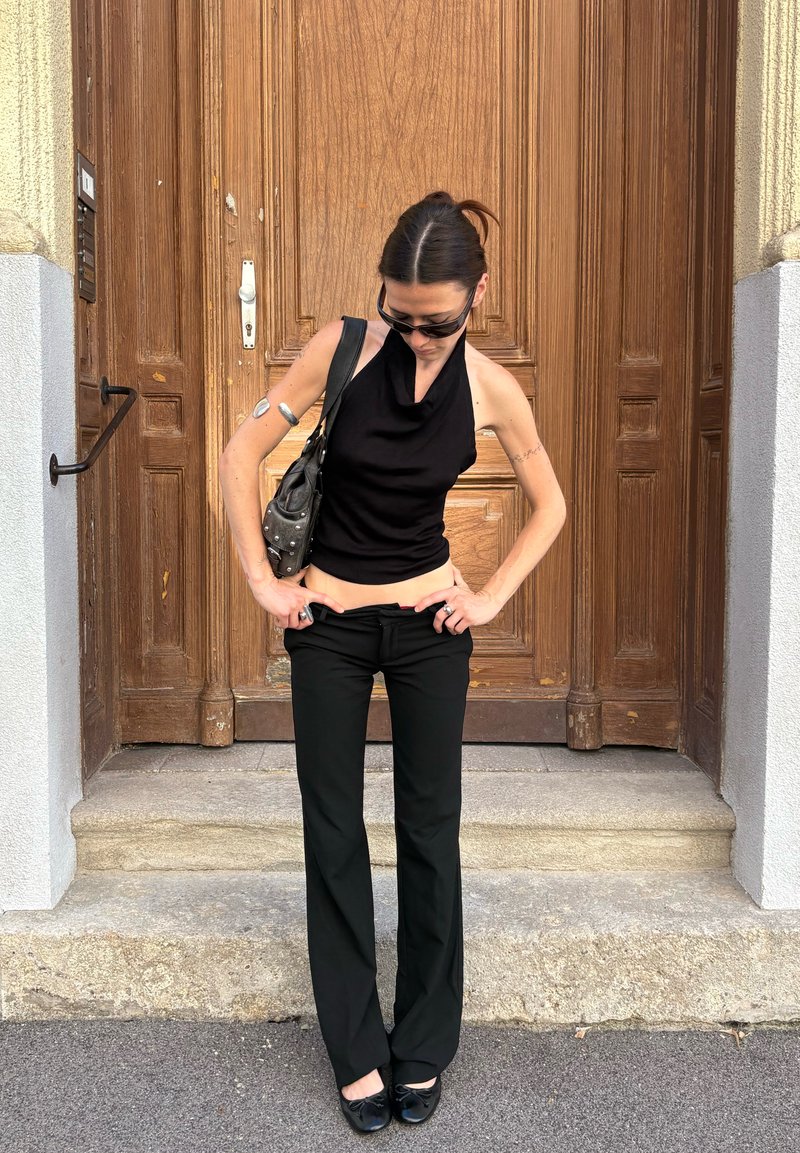 Femeie îmbrăcată într-un top negru fără mâneci și pantaloni își ajustează talia stând pe trepte de piatră în fața unei uși mari de lemn, poartă ochelari de soare și are o geantă.
