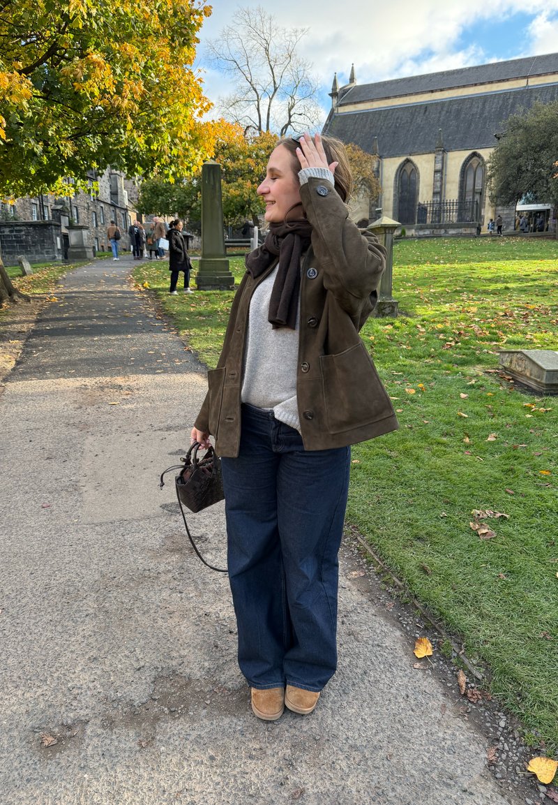 Bruine suède jas, grijze trui, donkerblauwe jeans met wijde pijpen, bruine laarzen en een kleine gevlochten tas. Buitenlocatie met bomen en een kerk.