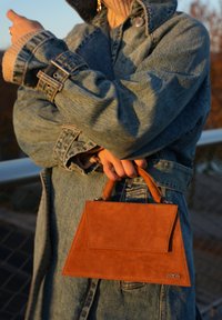 Rjava semiš torbica z obliko strukturiranega trikotnika, z ročajem na vrhu in sprednjo preklopno zaponko. Opremljena z detajlom metalnega logotipa.