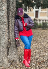 Jachetă scurtă de piele cu guler bleumarin, pulover roșu tricotat, blugi albaștri și cizme roșii de cauciuc. Stă lângă trunchiul unui copac texturat.