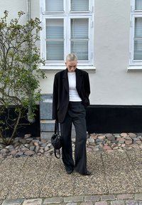 Zwarte oversized blazer, witte aansluitende top, zwarte wijde broek en een kleine ronde zwarte tas. Zwart-wit gestreepte schoenen op een stenen tegel.