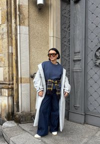 Długi szary płaszcz na granatowym swetrze, szerokie ciemne jeansy oraz czarno-złoty wiązany pas w kratę. Całość dopełniają duże prostokątne okulary przeciwsłoneczne.