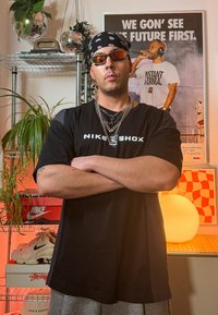 Svart T-shirt med korta ärmar och trycket "NIKE SHOX", lager på lager med silverkedjor, solglasögon, gråa shorts, med skor och en växt i bakgrunden.