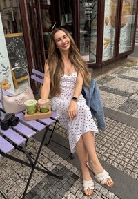 Biely šaty s bodkami, svetlomodrá denimová bunda, svetlobéžová kabelka, dva zelené smoothies v priesvitných pohároch, fialový drevený stôl, čierny fotoaparát.