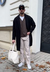 Mann in schwarzem Mantel, braunem Cardigan, weißen Hosen, weißen Sneakern, schwarzer Kappe und Sonnenbrille, der eine weiße Tragetasche auf einem Bürgersteig in der Stadt trägt.