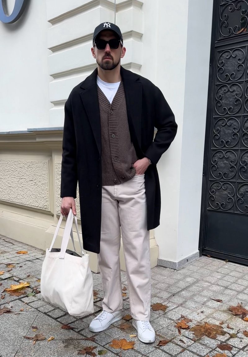 Mann in schwarzem Mantel, braunem Cardigan, weißen Hosen, weißen Sneakern, schwarzer Kappe und Sonnenbrille, der eine weiße Tragetasche auf einem Bürgersteig in der Stadt trägt.