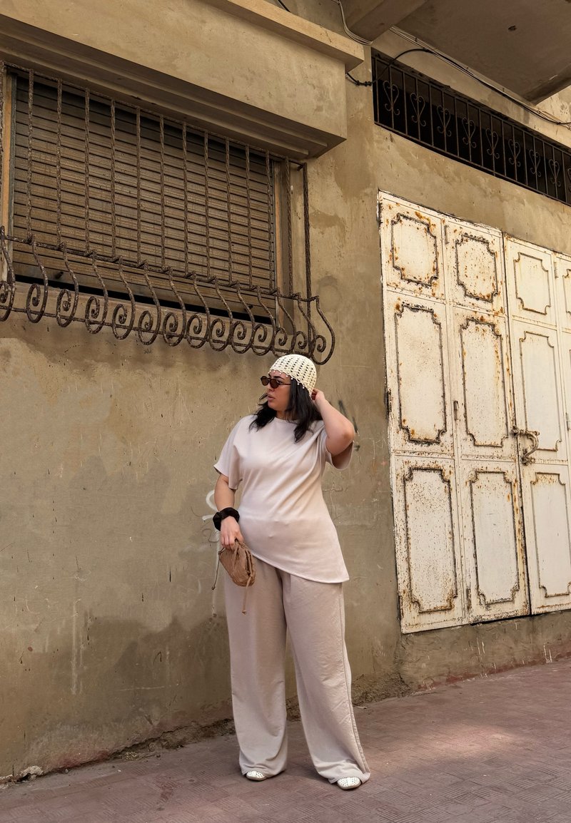 Woman in beige outfit, crocheted hat, holding a small bag, standing by a weathered wall with rusted white metal doors and barred window.