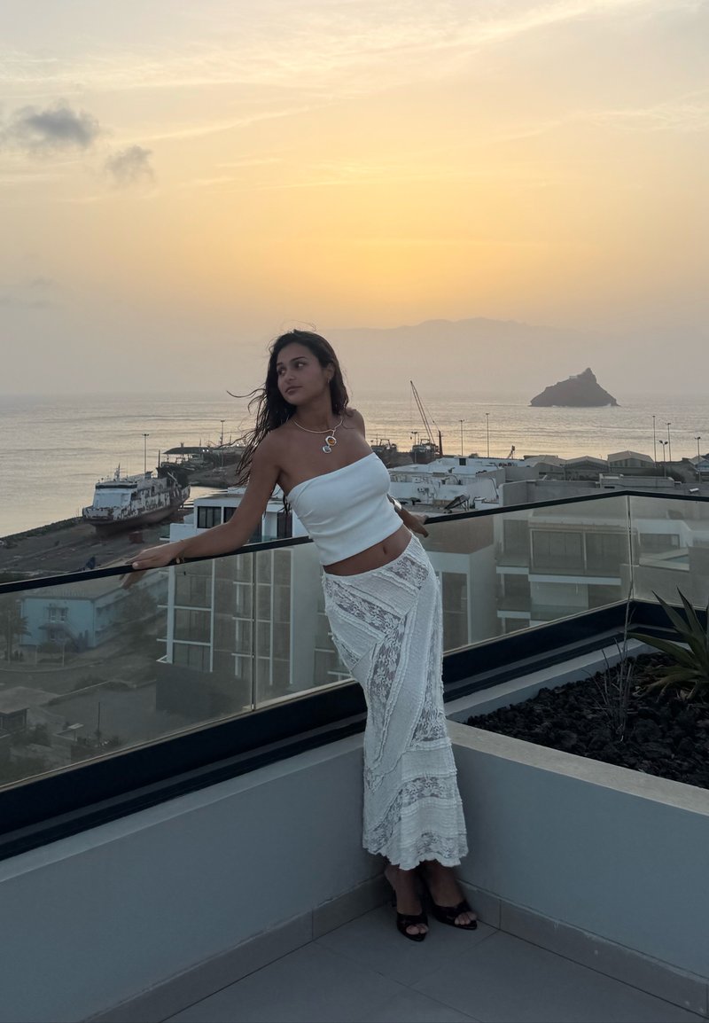Femme en haut blanc et longue jupe en dentelle s’appuie sur la rambarde en verre du balcon au coucher du soleil avec océan, bâtiments et bateau en arrière-plan.
