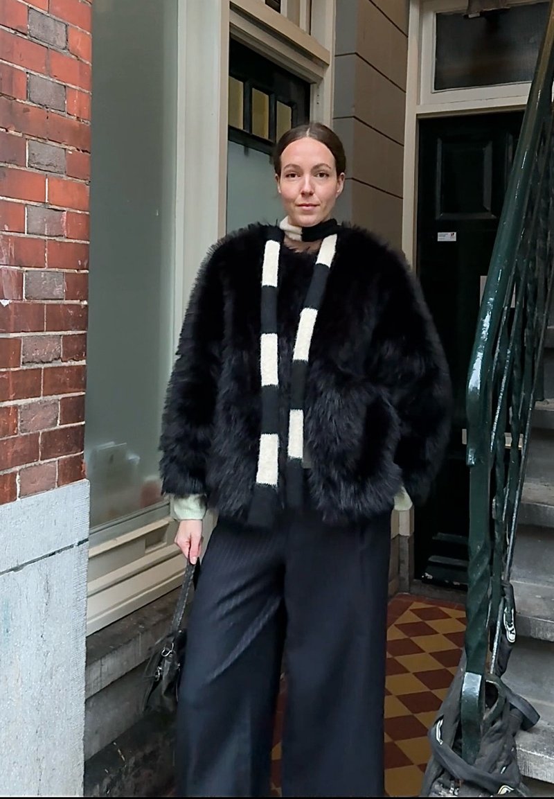 Femme portant un manteau en fausse fourrure noire, une écharpe noire et blanche, et un pantalon large noir, debout près d'un mur en briques et d'un escalier en métal.