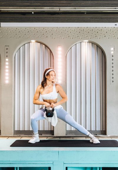 Mujer atlética con una camiseta blanca y leggings azul claro realizando una zancada con una kettlebell sobre una esterilla negra, con un elegante fondo iluminado.