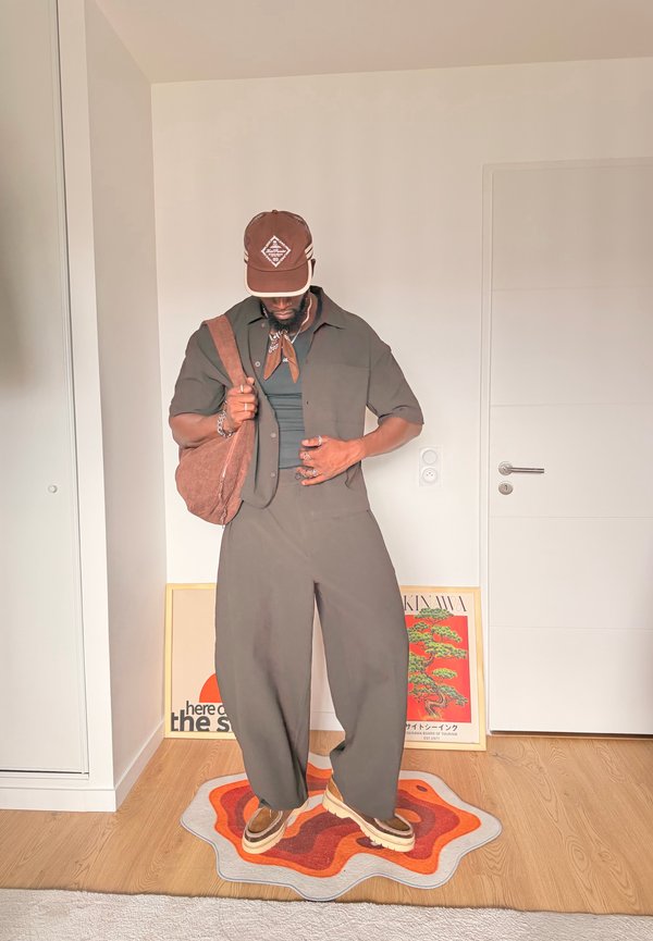 Homme portant une casquette marron, une tenue vert foncé, des chaussures marron et portant un sac à épaule marron, debout sur un tapis abstrait multicolore dans une pièce avec des affiches.