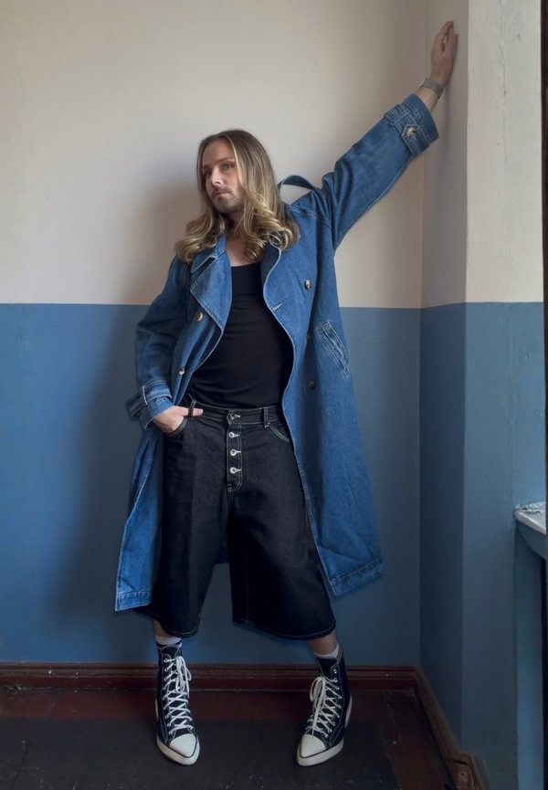 Persona con cabello largo y rubio se apoya en la esquina de una pared vistiendo un abrigo azul de mezclilla, camiseta negra, pantalones cortos largos negros y zapatillas negras altas.