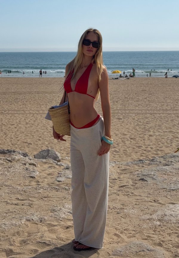 Frau in rotem Bikini-Oberteil und weißer Hose mit Sonnenbrille und Strohtasche steht an einem sandigen Strand in der Nähe des Ozeans an einem sonnigen Tag.