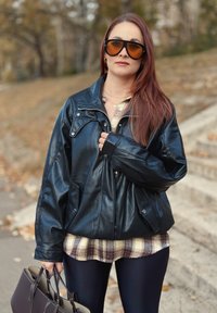 Sort læderjakke med en løs pasform, lynlås og sidelommer, lagdelt over en ternet skjorte. Båret med mørke leggings og en håndtaske.