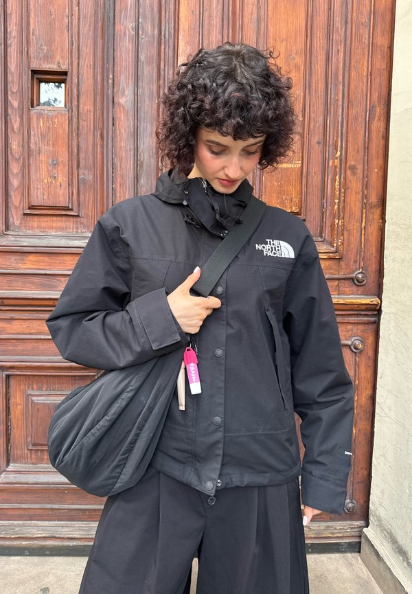 Une personne portant une veste noire North Face regarde vers le bas tout en tenant un grand sac noir en bandoulière devant une porte en bois.