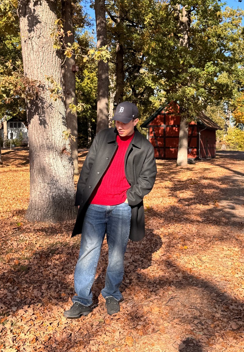 Sort frakke, rød strikket sweater, blå jeans og sorte sko. Står på orangebrune blade med træer og en rød lade i baggrunden.